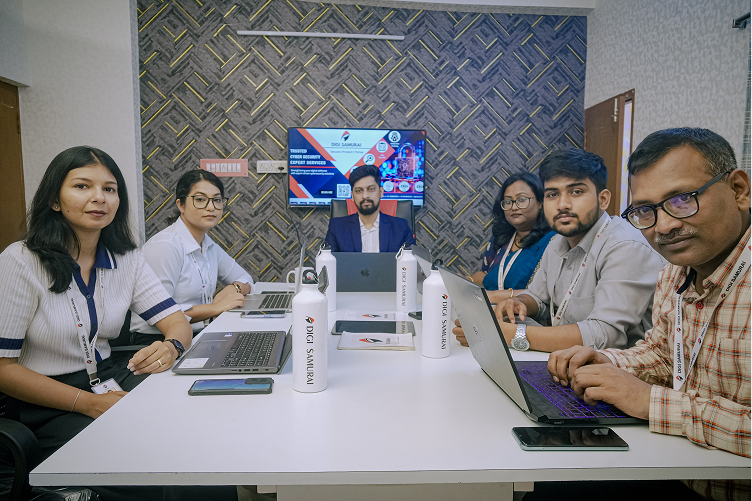 Team of six people sitting around a white table with laptops and a TV screen on the wall in a meeting room